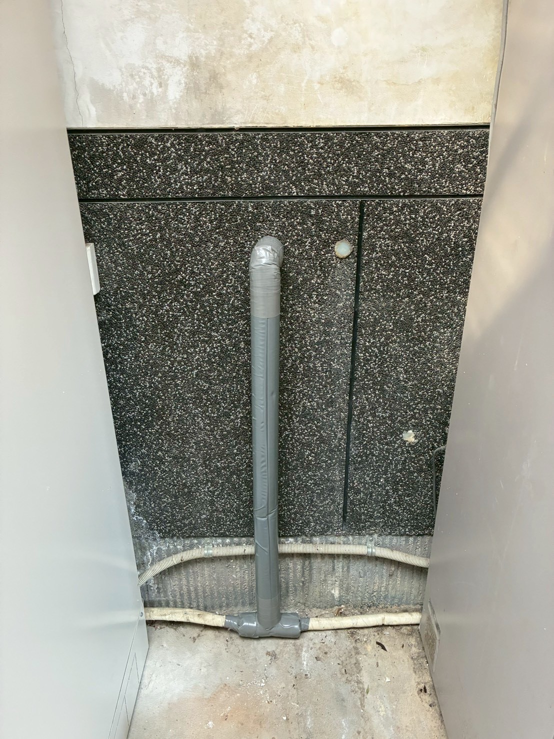 屋外の黒いパネルにグレーの保温管が縦に通っている施工前の状態